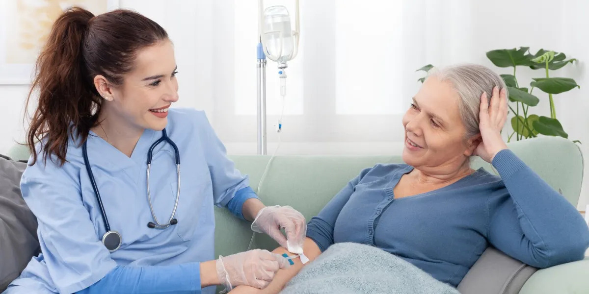 young smiling nurse or doctor help sick senior elderly woman patient in receiving iv drip infusion and vitamin injection therapy sitting on sofa in medical clinic or at home health care concept