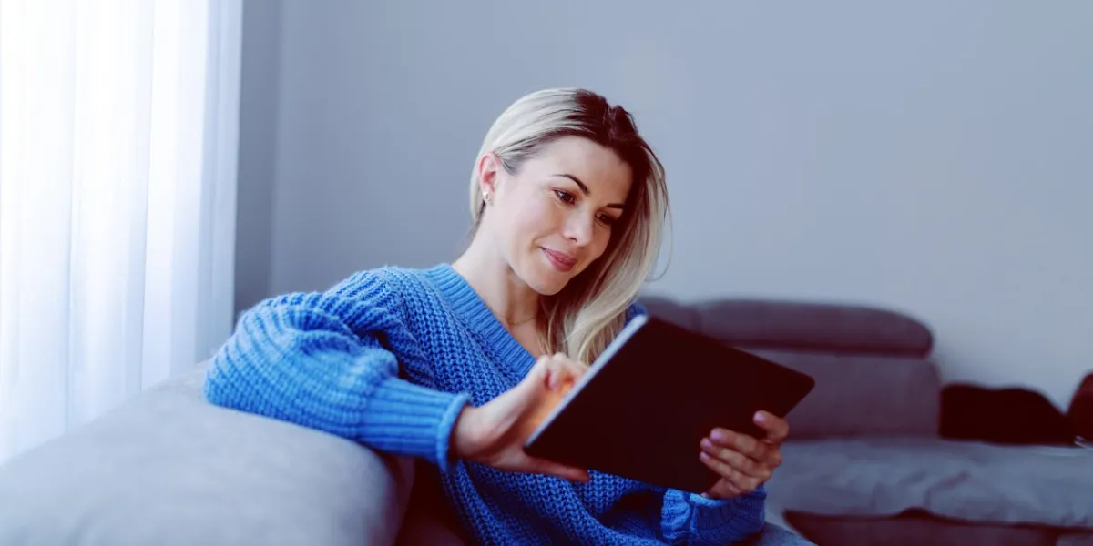 beautiful caucasian smiling blonde in blue sweater and jeans sitting on sofa in living room and using tablet apartment interior