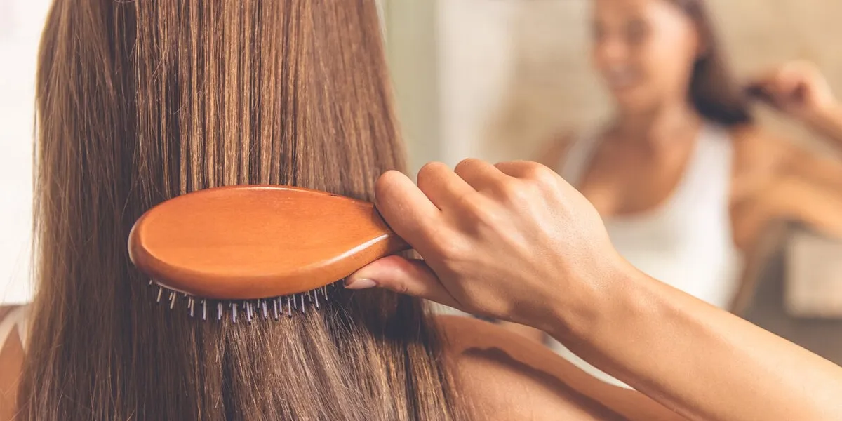 back view of beautiful young woman in white undershirt combing her hair and smiling while looking into the mirror in bathroom