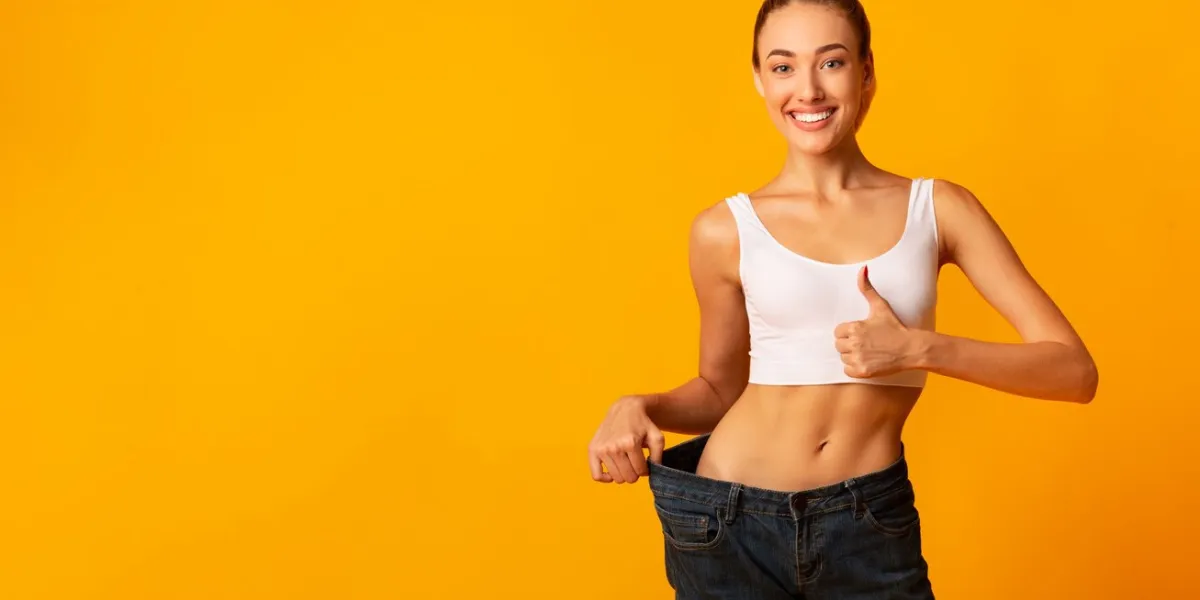 weight loss girl in oversize jeans gesturing thumbs up smiling at camera standing on yellow studio background copy space