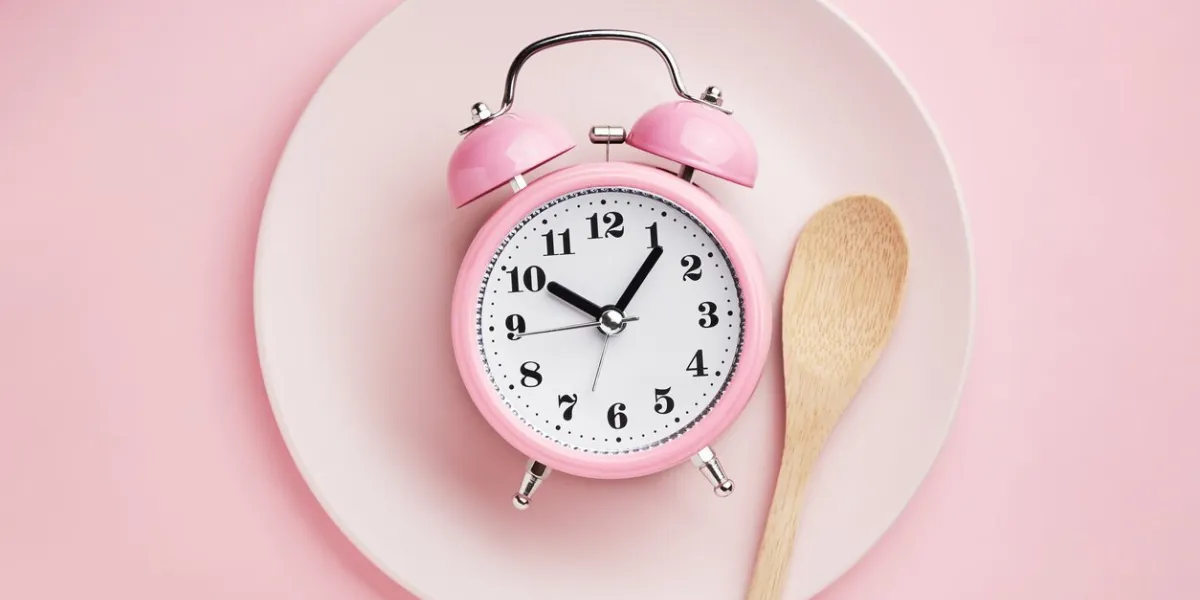 alarm clock and wooden spoon on empty pink plate concept of intermittent fasting, lunchtime, diet and weight loss