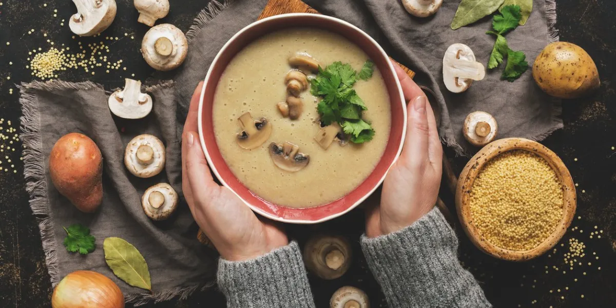 women hands in a gray sweater holding a bowl of cream of mushroom soup hot winter soup on a dark rustic background top view, flat lay