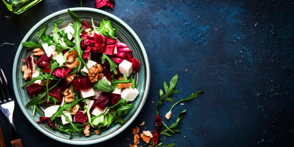 arugula, beet and cheese salad with fresh radicchio and walnuts on plate with fork, dressing and spices on blue kitchen table background, place for text, top view