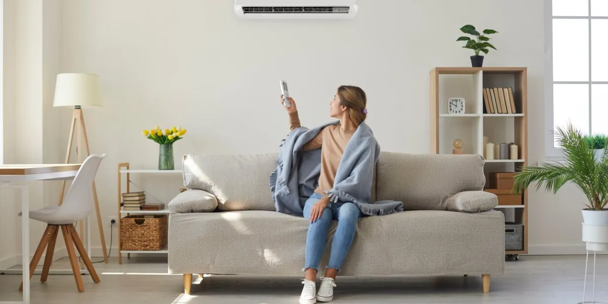 woman who's sitting on sofa under warm plaid in living room switches off her air conditioner on wall young girl adjusting modern ac system, regulating temperature and enjoying cool fresh air at home