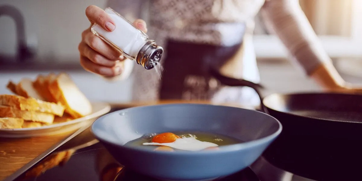 close up of caucasian woman adding salt in sunny side up eggs while standing in kitchen next to stove
