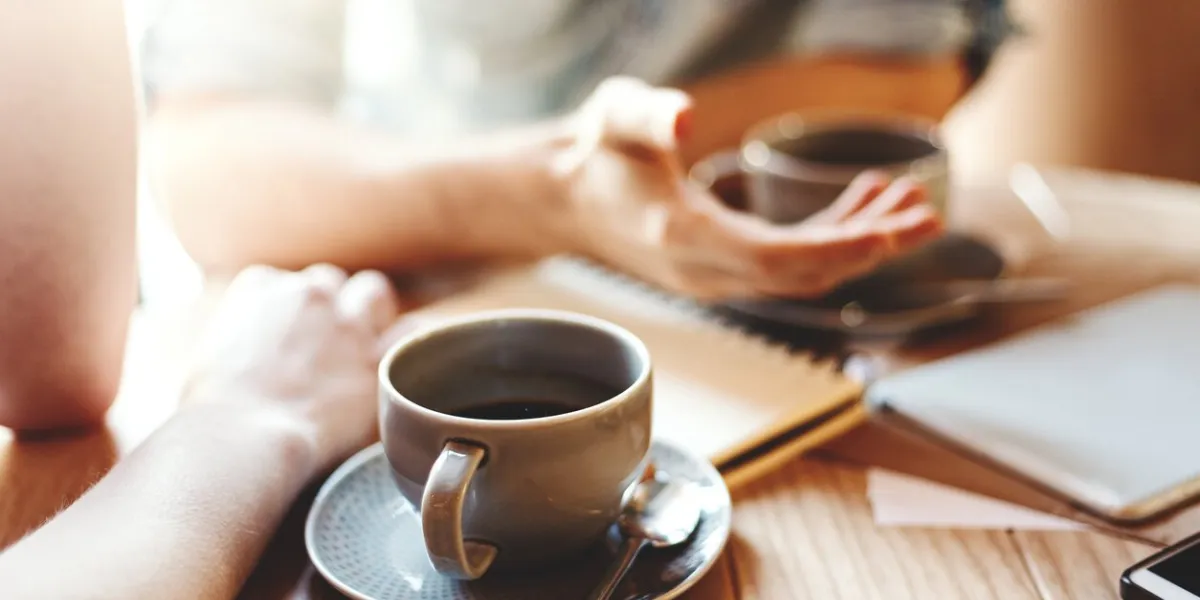 friends talking at cafe table during coffee break unrecognizable male and female colleagues discussing business issues, focus on coffee cup with saucer and teaspoon