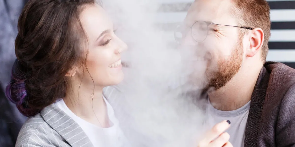 couple vaping young man and woman blowing smoke at black and white striped studio background relationship and vape addiction concept with copy space close-up portrait