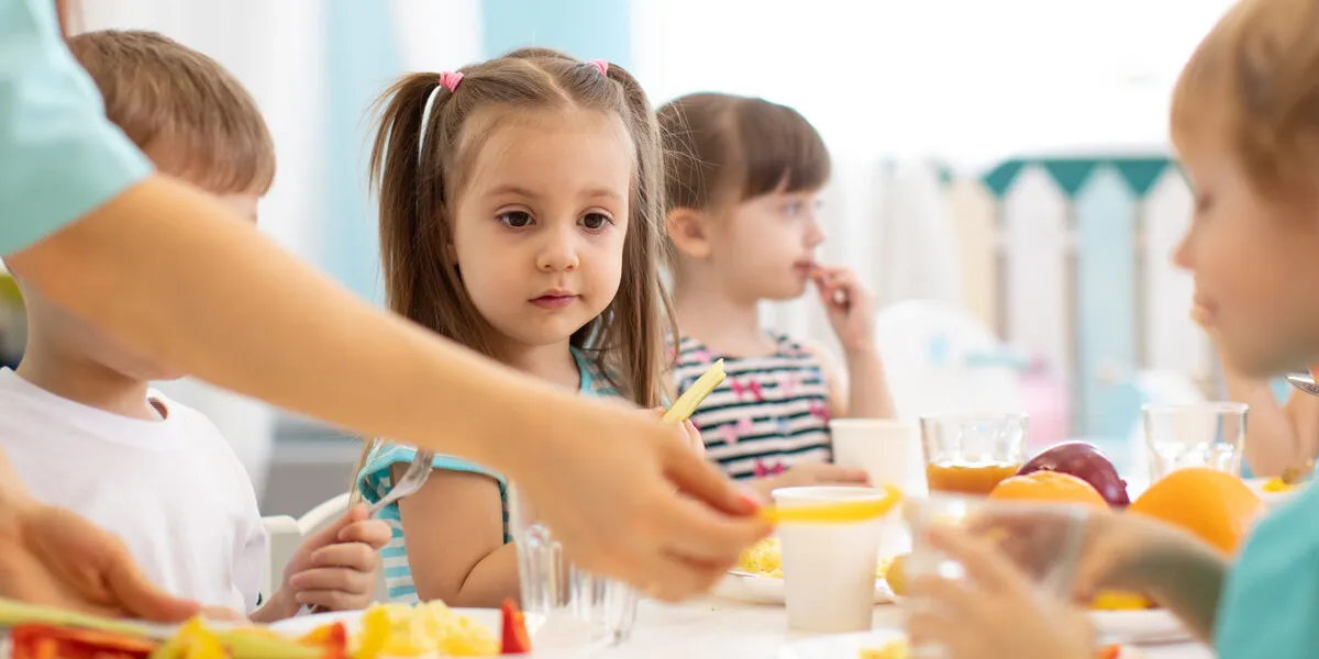 kids have a dinner in preschool little boys and girls from the group of children sit at table with lunch and eat appetizing children with caregiver in day care centre