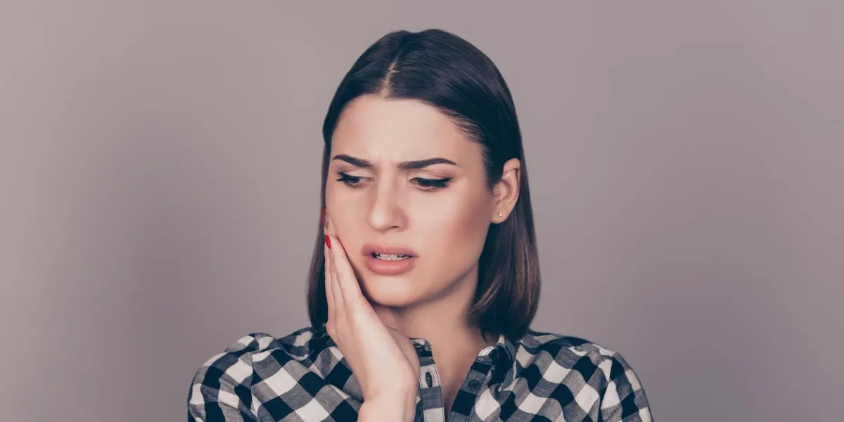a horizontal photo of a young worried woman in checkered shirt suffering from strong toothache and touching her cheek