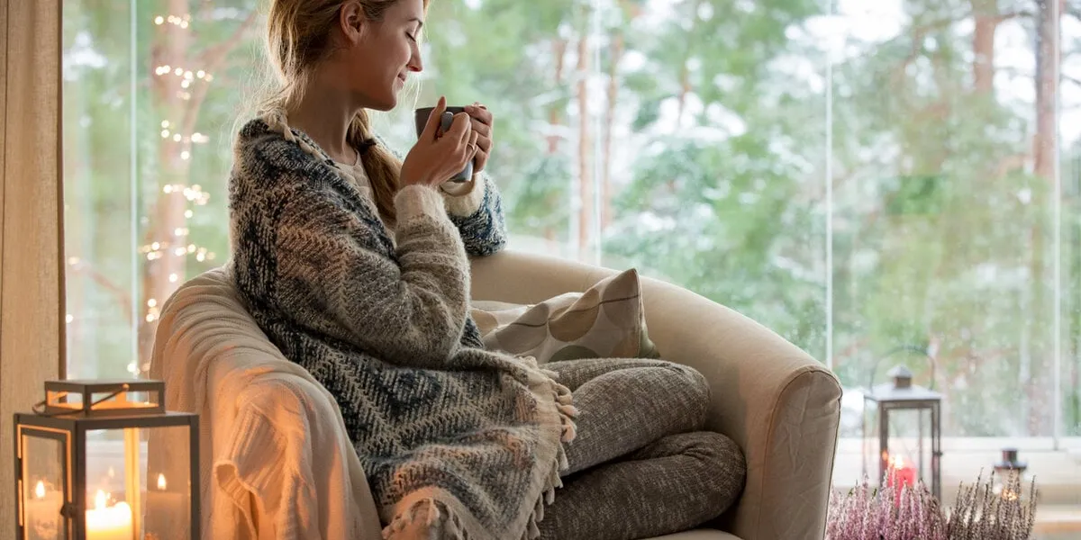 young beautiful woman sitting home in the chair by the window with cup of hot coffee wearing knitted warm sweater cozy room decorated with lanterns and candles