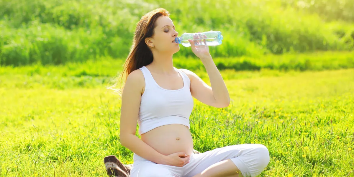 grossesse, sport et santé mode de vie - jeune femme enceinte l'eau potable d'une bouteille assis sur l'herbe dans la journée d'été ensoleillée