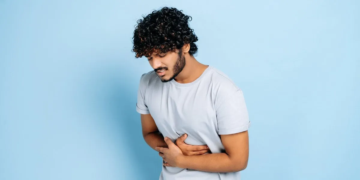 stomach ache indian guy with in casual clothes hunching and holding his belly, suffers from pain in the abdomen or stomach, got poisoned, need treatment, stands on isolated blue background
