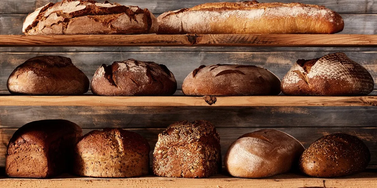 bunch of assorted freshly baked bread loaves with different shapes and baguettes placed on shelves against wooden background in light studio