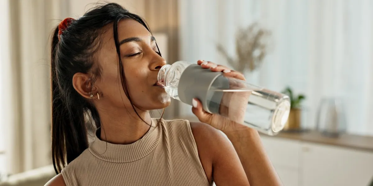 drinking water, yoga or indian woman in home with health, fitness or wellness for natural hydration thirsty female person, tired or healthy girl with liquid bottle after pilates to detox or relax