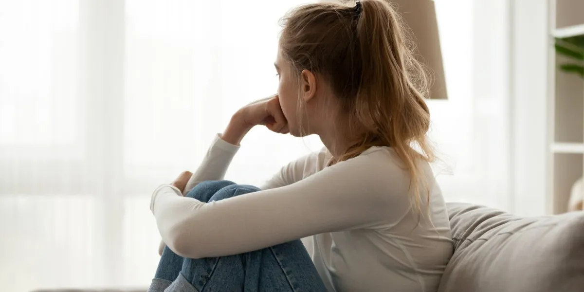 side view young woman looking away at window sitting on couch at home frustrated confused female feels unhappy problem in personal life quarrel break up with boyfriend or unexpected pregnancy concept