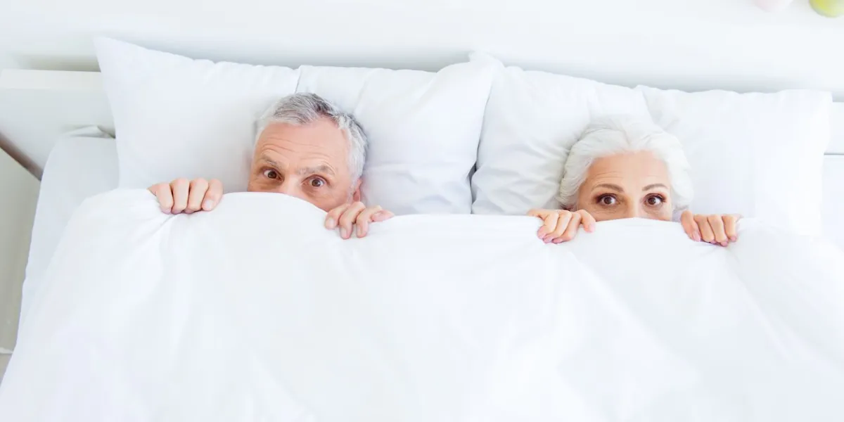 high angle top view surprised funny couple gray hair people in pajama, sleep, wear, sleepwear, nightwear with big eyed lying in the bed linen, sheets hiding themselves under blanket
