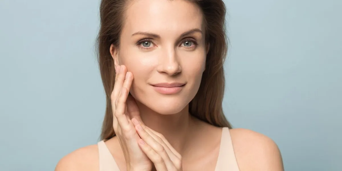 close up portrait of beautiful young woman isolated on blue studio background touch clean healthy skin, pretty millennial female model look at camera satisfied with moisturized hydrated face treatment