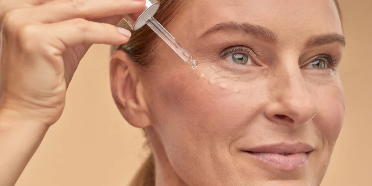 smiling mature woman with fresh skin using eye cream, isolated on beige background beauty treatment and wellness concept