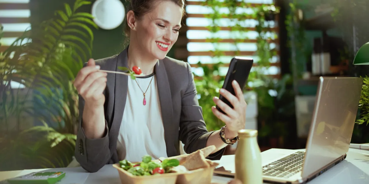 sustainable workplace smiling modern female worker in a grey business suit in modern green office with laptop and smartphone eating salad