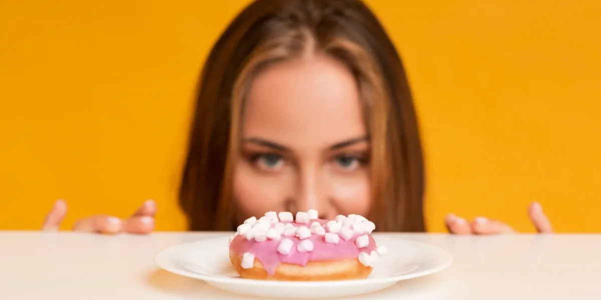 im on diet hungry woman peeking out of behind table and looking at donut with appetite, yellow studio background with empty space, selective focus