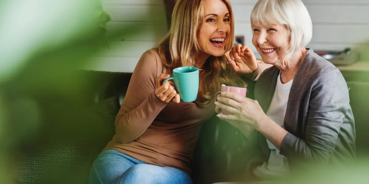 happy senior mother with adult daughter sitting on couch and holding cups with coffee or tea at home togetherness concept