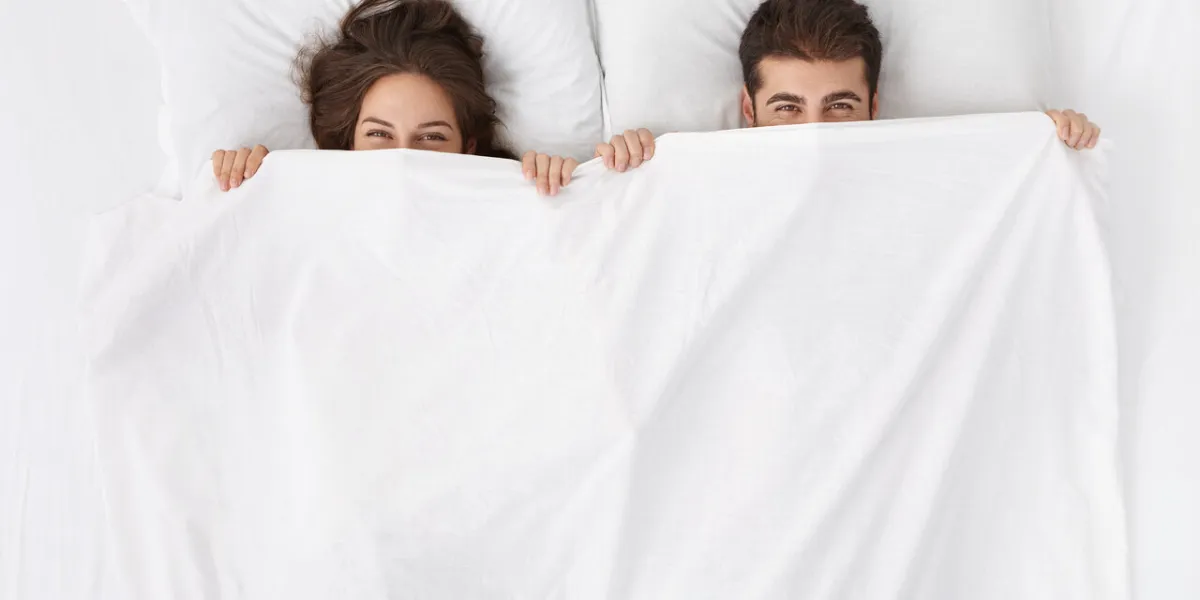 funny married couple lying in bed and hiding under white blanket, looking at camera with eyes full of joy attractive caucasian man and woman having fun in bedroom love and happiness concept