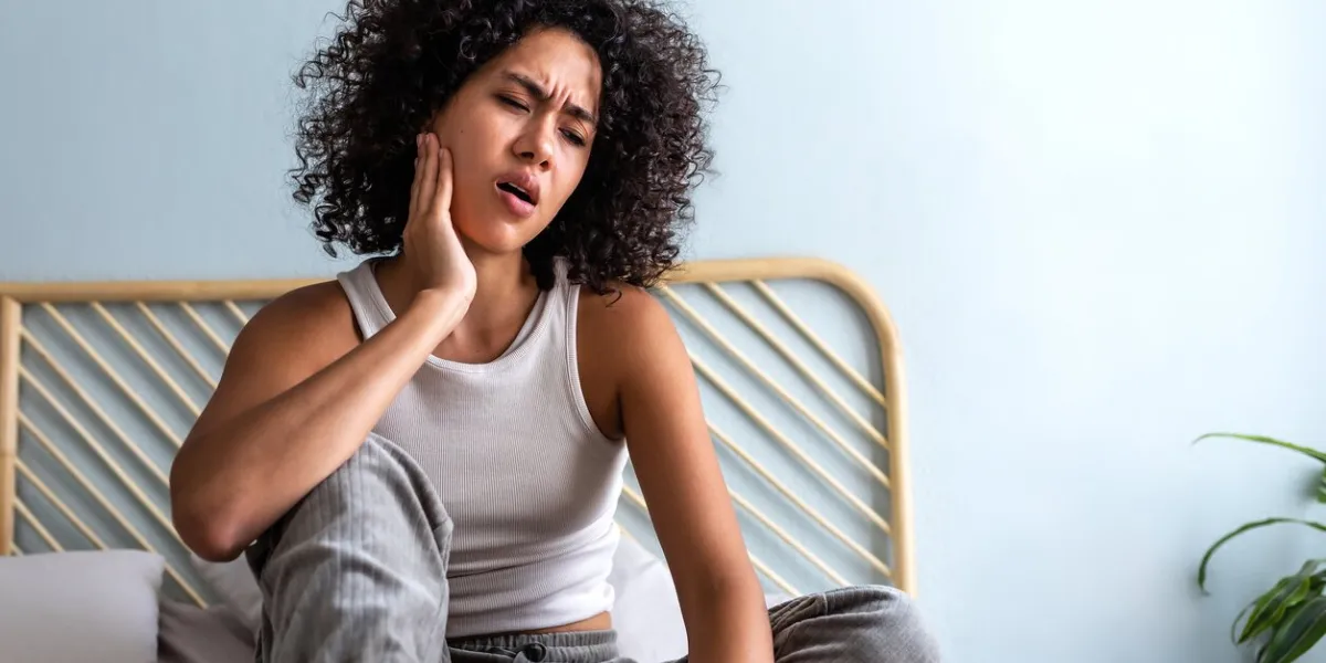 young latina female with curly hair touching cheek in pain having toothache sitting on bed in cozy bedroom at home body concept
