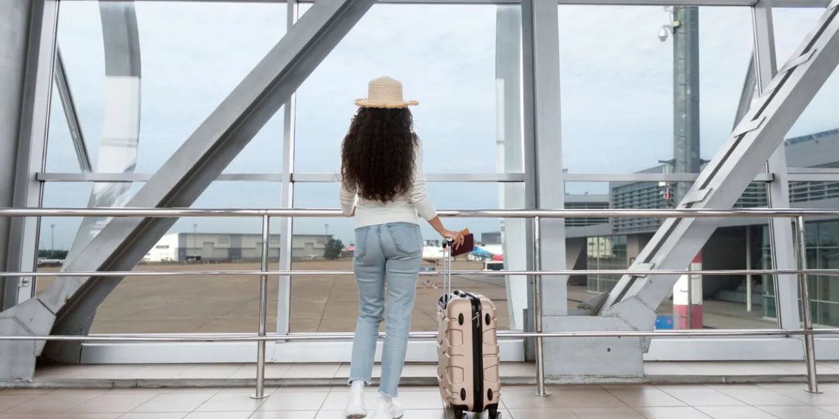 air travels concept rear view of female traveller in wicker hat waiting in airport, unrecognizable young woman standing in terminal with suitcase and looking from window, ready for trip, copy space