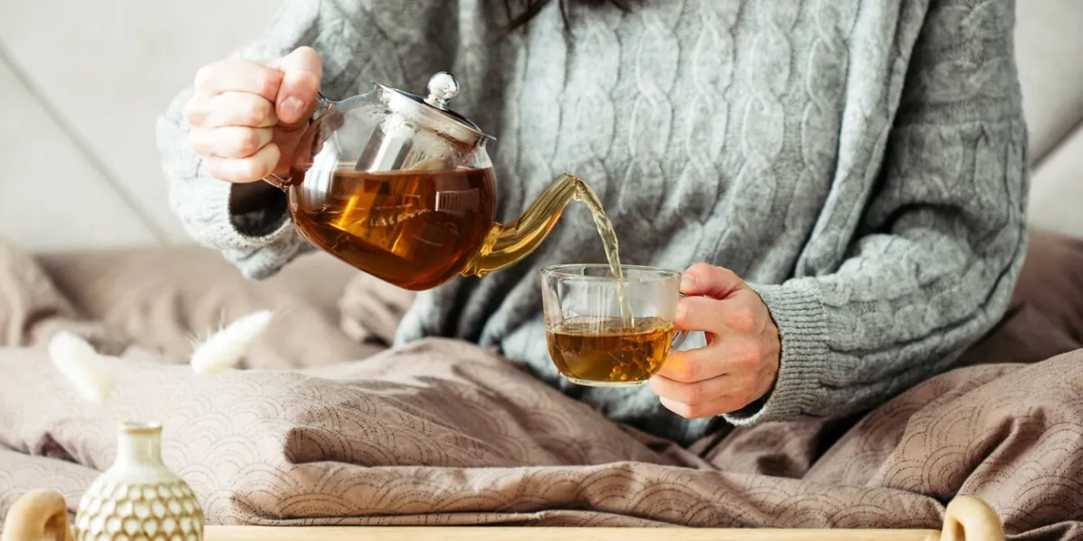 woman dressed in knitted sweater pours hot tea from glass teapot into mug while sitting in bed morning breakfast in cozy home bedroom interior hygge, warm, autumn concept