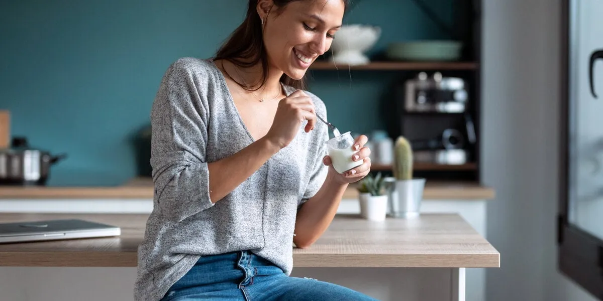 shot of smiling young woman eating yogurt while sitting on stool in the kitchen at home