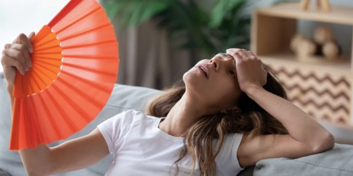 overheated woman sitting on couch, waving orange paper fan close up, girl feeling unwell, suffering from heating at home, feeling discomfort, hot summer weather or fever, sitting on couch alone