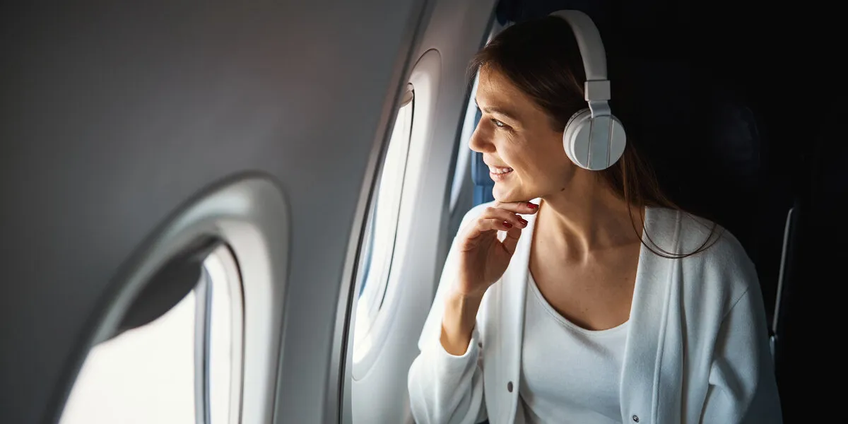 positive young woman in headphones smiling while looking outside of the airplane website banner