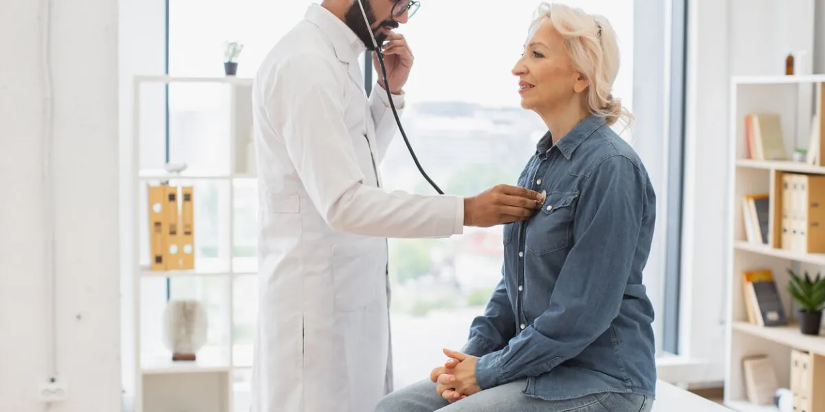 side view of focused adult man in white coat using stethoscope while elderly woman sitting on exam couch general practitioner examining heartbeat while providing regular checkup in clinic