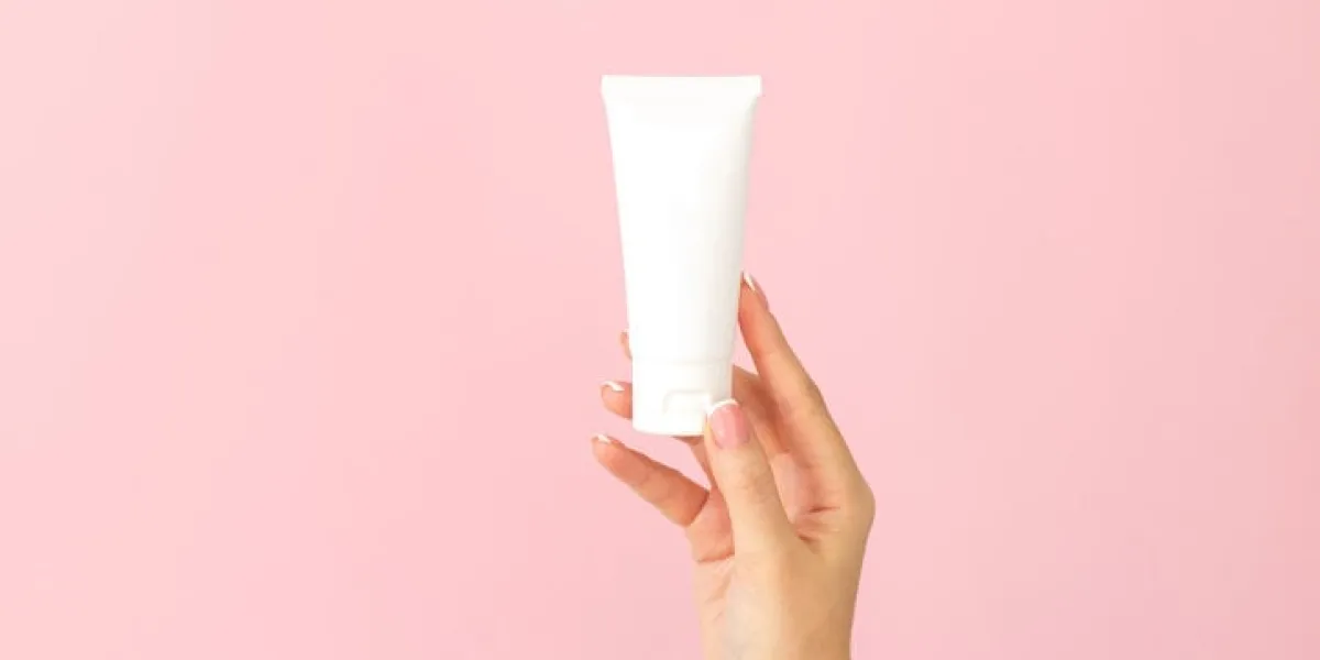 woman holding cream tube and applying moisturizer lotion on her beautiful hands for soft and healthy skin on pink background