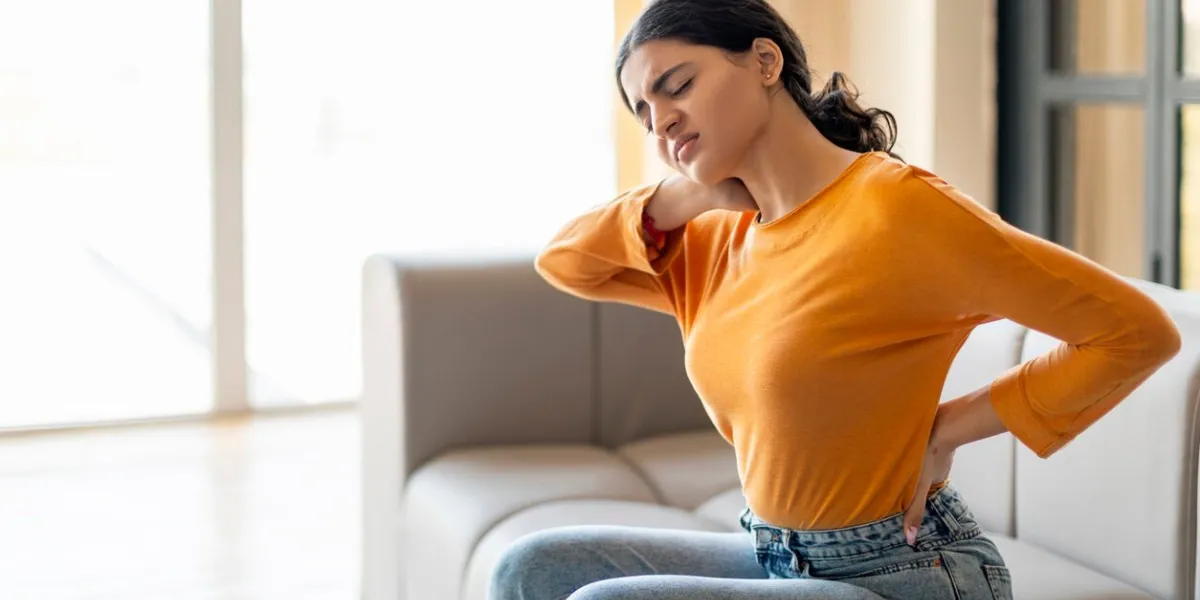 young indian woman suffering neck and back pain at home, sick eastern female rubbing painful sore zones and frowning, massaging aching areas while sitting on couch in living room, copy space