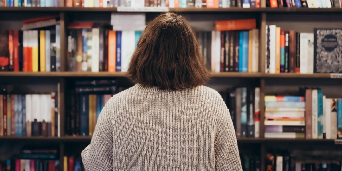 young brunette woman choosing a book in a bookstore, back view confused young woman seen from behind looking a the bookshelves thinking what book to buy at the bookstore