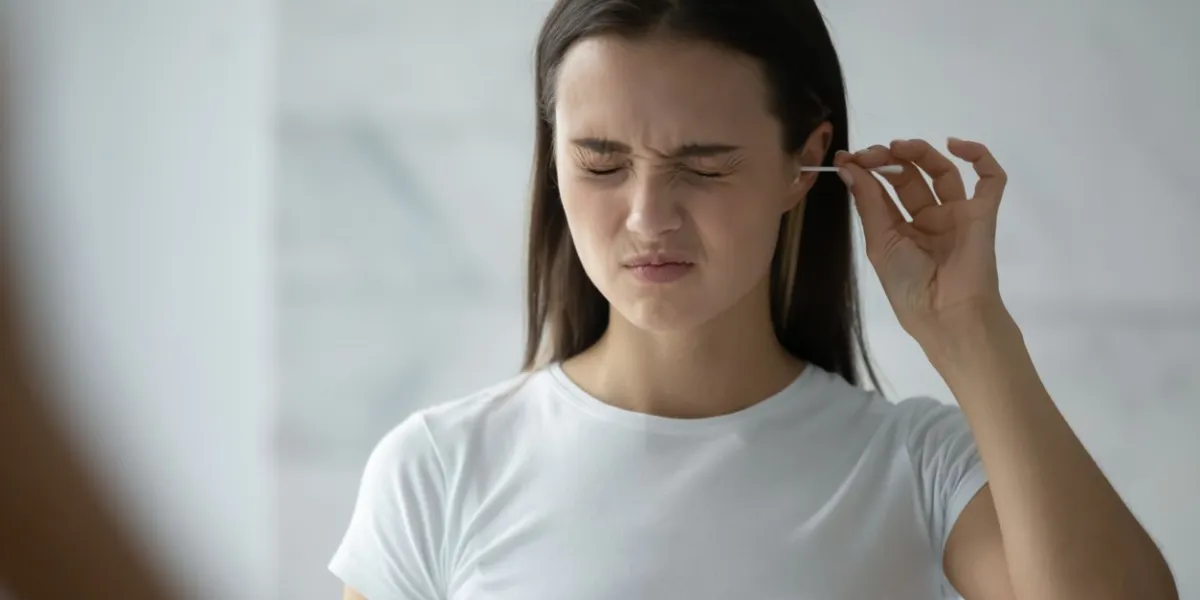 young woman stand in bathroom look in mirror clean ears with cotton bud, millennial girl feel pain cleansing using stick or swab after shower in home bath, hygiene, daily routine concept