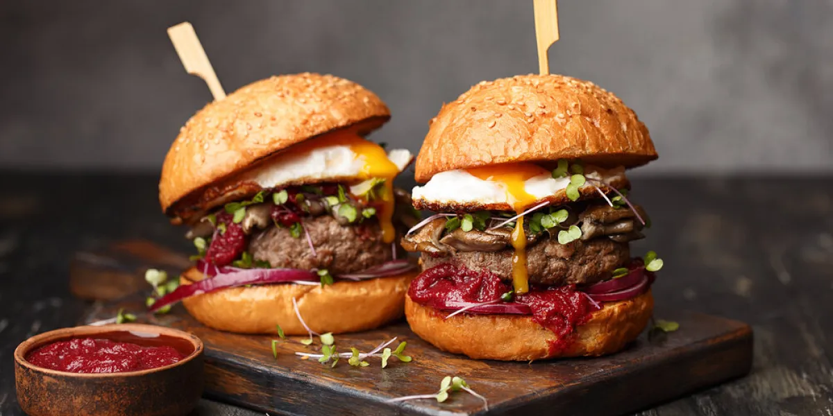 two homemade beef burgers with mushrooms, micro greens, red onion, fried eggs and beet sauce on wooden cutting board side view, close up