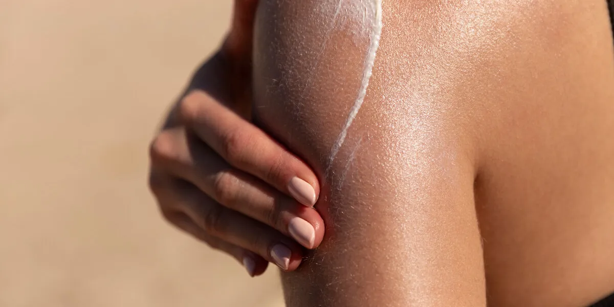 young woman applying sun cream or sunscreen on her tanned shoulder to protect her skin from the sun shot on a sunny day with blurry sand in the background