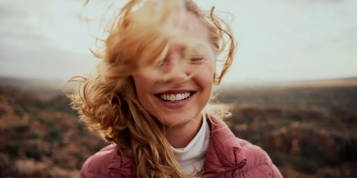 portrait of young smiling woman face partially covered with flying hair in windy day standing at mountain - carefree woman