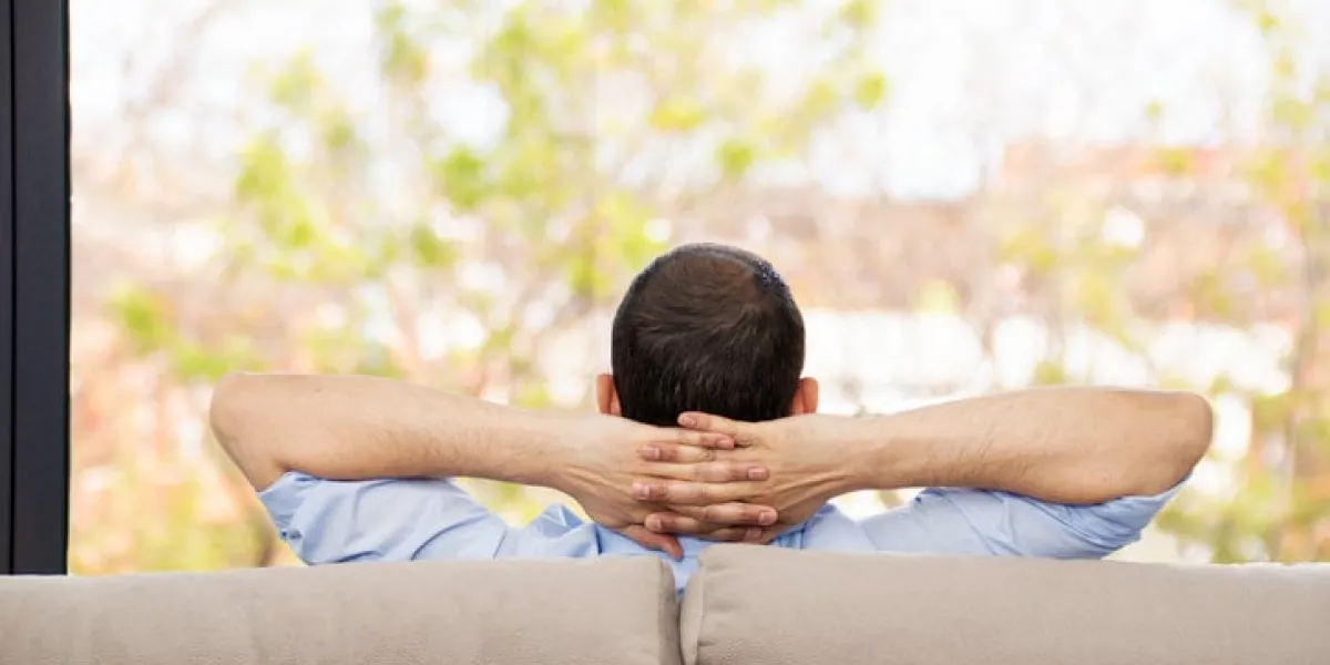 rear view of a single man relaxed on a couch at home and looking the green background outdoors through the window in the living room at home
