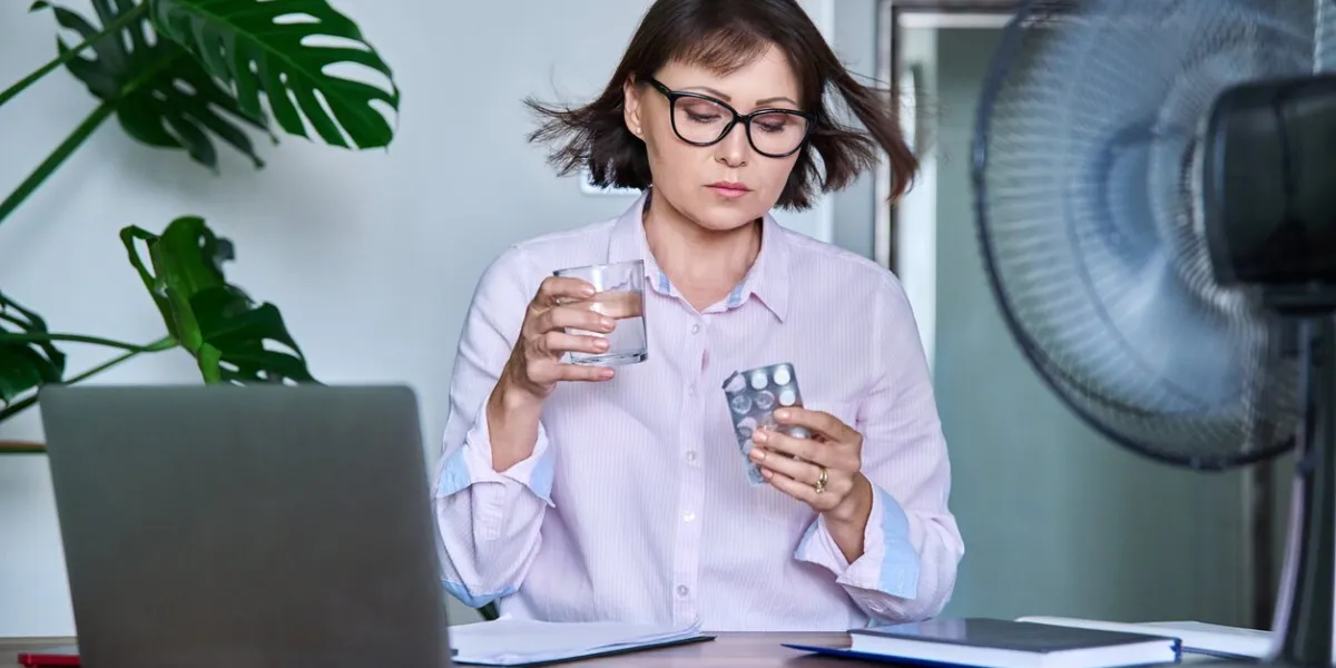mature woman with glass of water blister of pills taking medicine vitamins to relieve menopause symptoms, sitting at desk at workplace with computer laptop