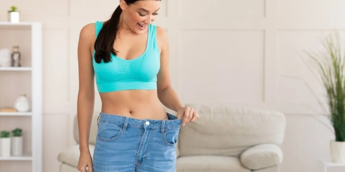slimming excited woman wearing too big jeans after weight loss standing at home empty space for text