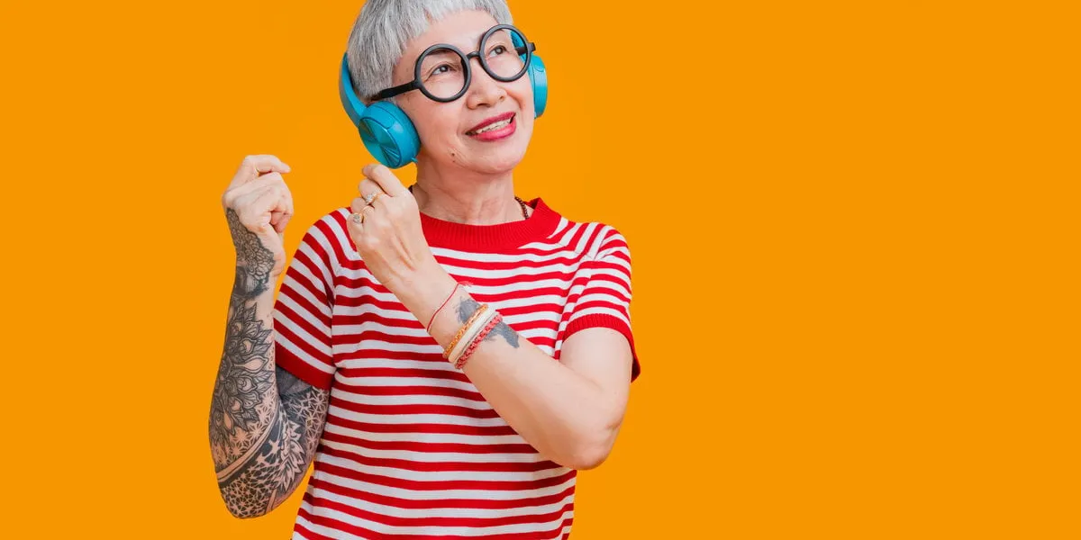 cheerful elderly gray-haired asian female woman with tattoo casual cloth listening music with headphones keeping eyes closed isolated on pastel yellow color background studio portrait