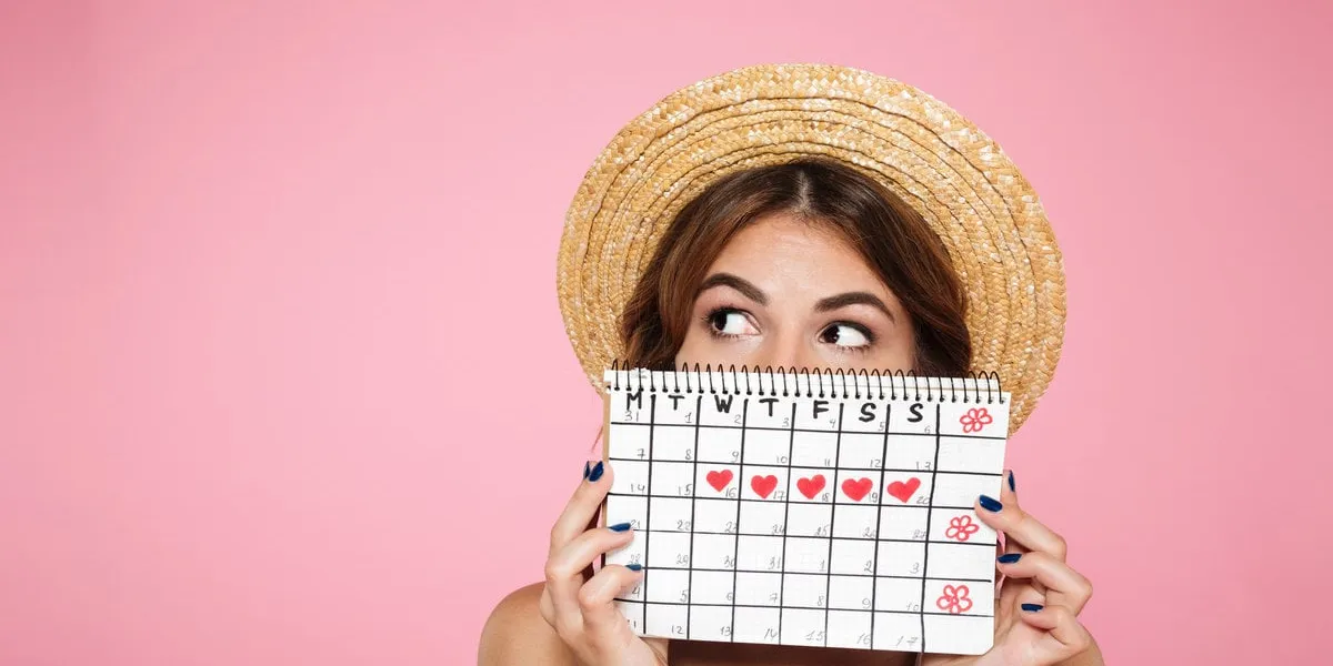 portrait of a funny young girl in summer hat hiding behind a periods calendar and looking away at copy space isolated over pink background