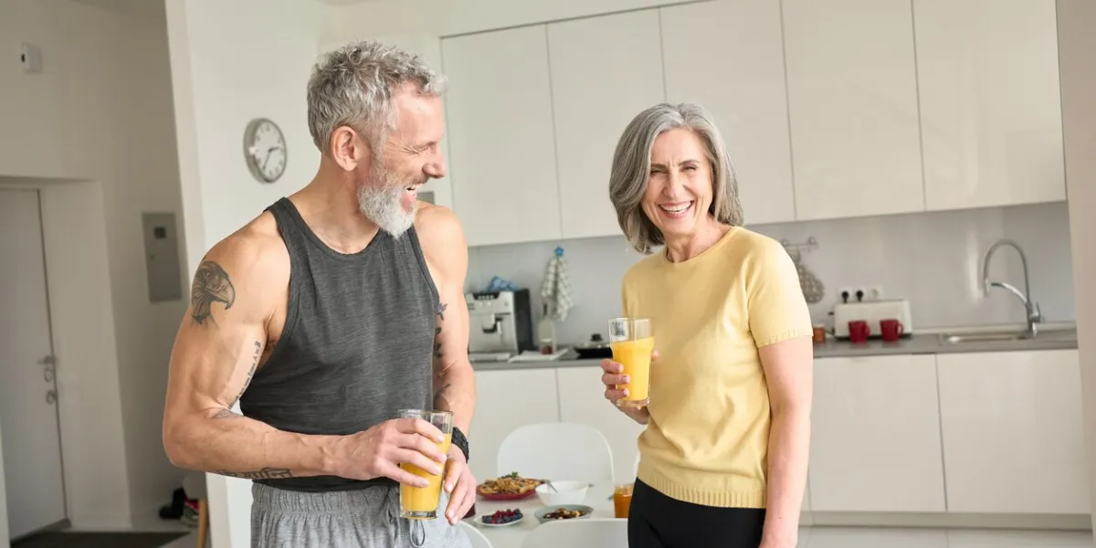 healthy sporty happy senior adults family couple drinking juice and laughing at home cheerful fit mid age older husband and wife enjoying talking, having fun standing in kitchen in the morning