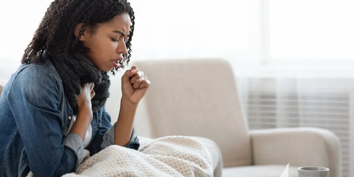 risk of coronavirus sick black woman coughing hard at home, sitting on couch wearing scarf and covered with blanket, side view with copy space