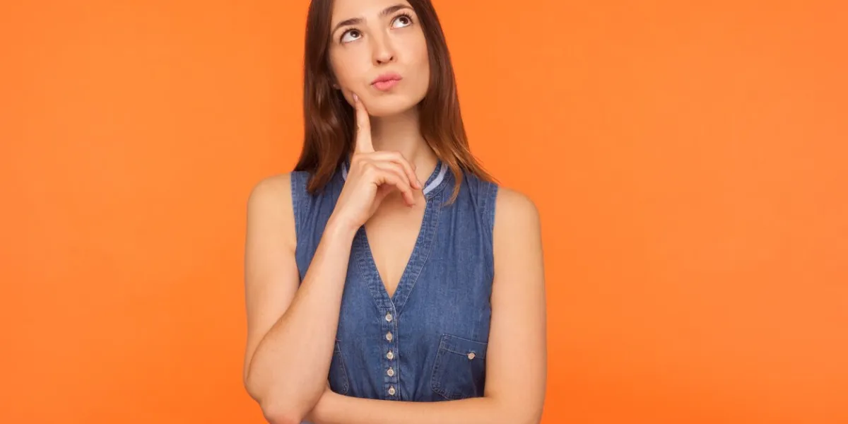 pensive brunette woman in denim dress looking up with thoughtful doubtful expression, pondering serious difficult idea, imagination and vision in mind indoor studio shot isolated on orange background