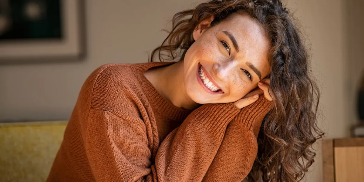 happy young woman sitting on sofa at home and looking at camera portrait of comfortable woman in winter clothes relaxing on armchair portrait of beautiful girl smiling and relaxing during autumn