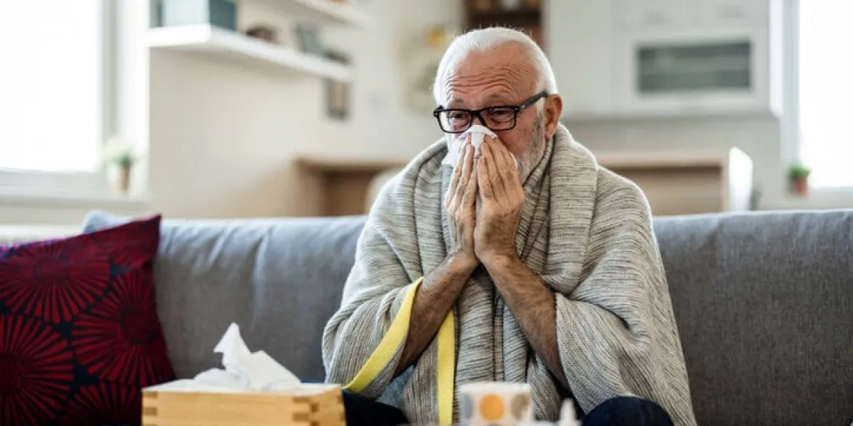 cold exhausted senior man with flu wrapped in a warm blanket blowing his nose with a tissue in the livingroom man with a cold lying in sofa holding tissues